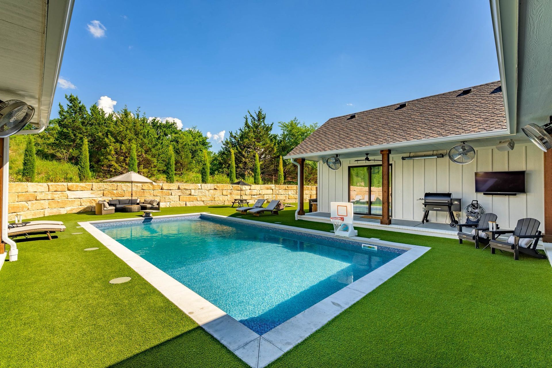 Backyard with pool, patio, and house under a sunny sky. Green turf, stone wall, trees.