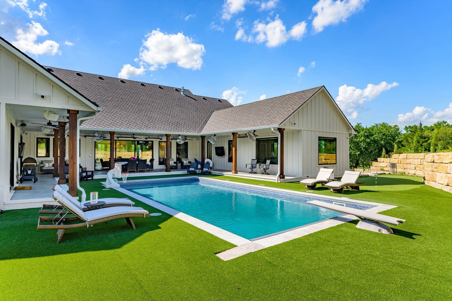Backyard with pool and white farmhouse, lounge chairs, green grass and blue sky.