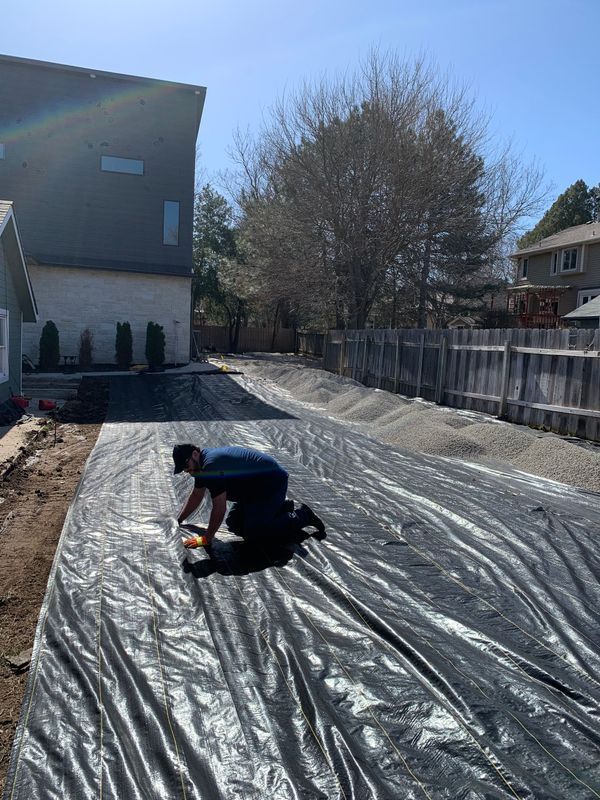 Man kneels on black tarp, cutting it in a backyard with a house and fence. Sunny day.
