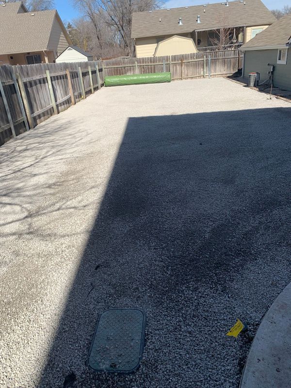 Gravel-covered backyard with a fence and house in the background. Sunlit area with partial shadow.