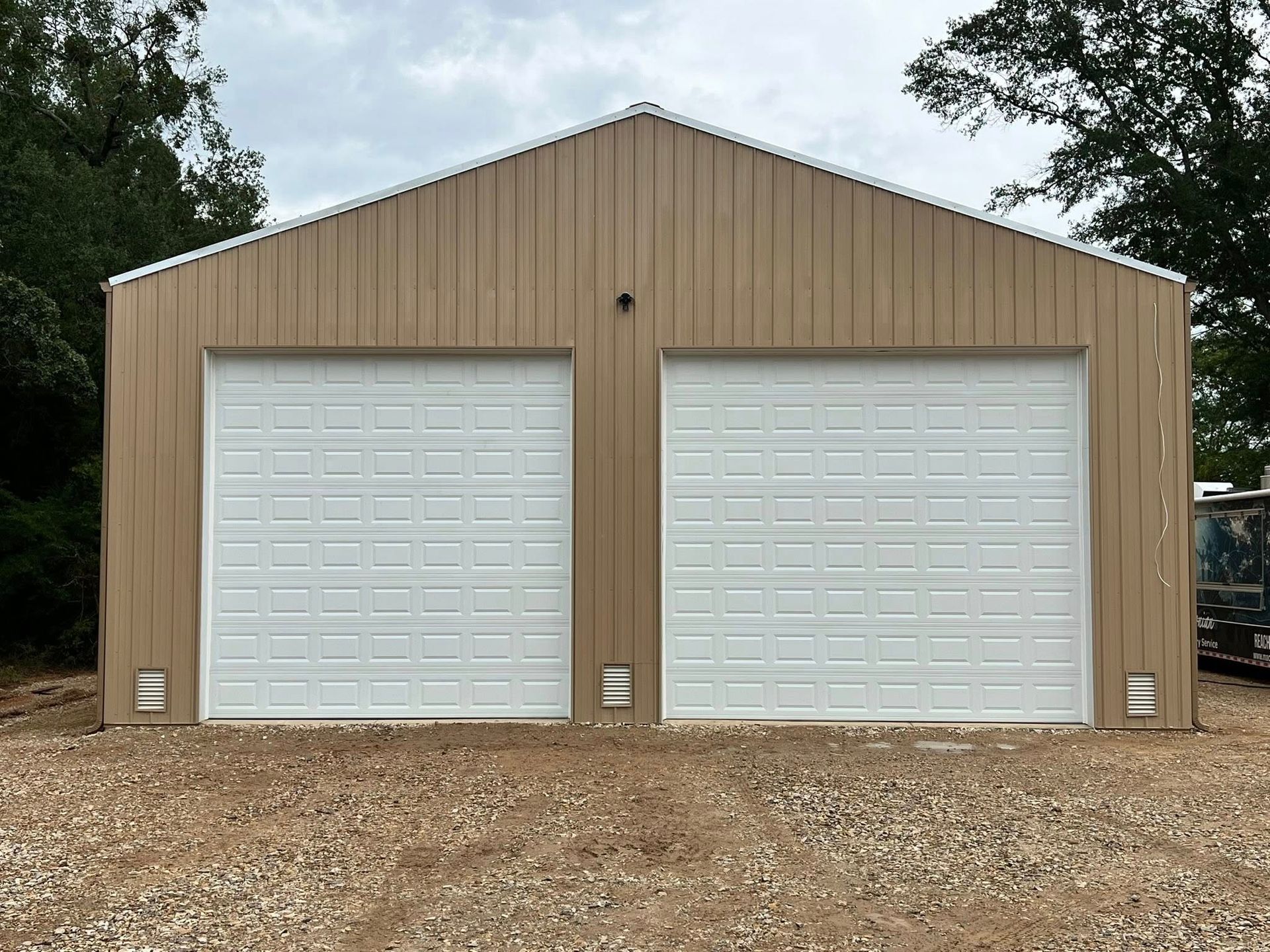 Tan metal two-car garage with white garage doors, set on a gravel driveway.