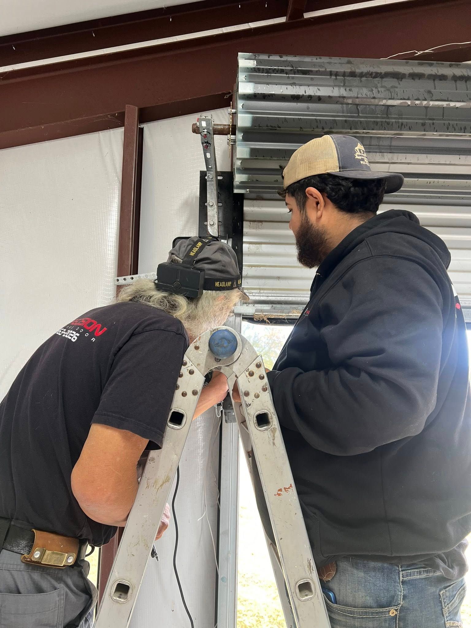 Two workers installing a metal roll-up door in a building. One on ladder, other assisting.