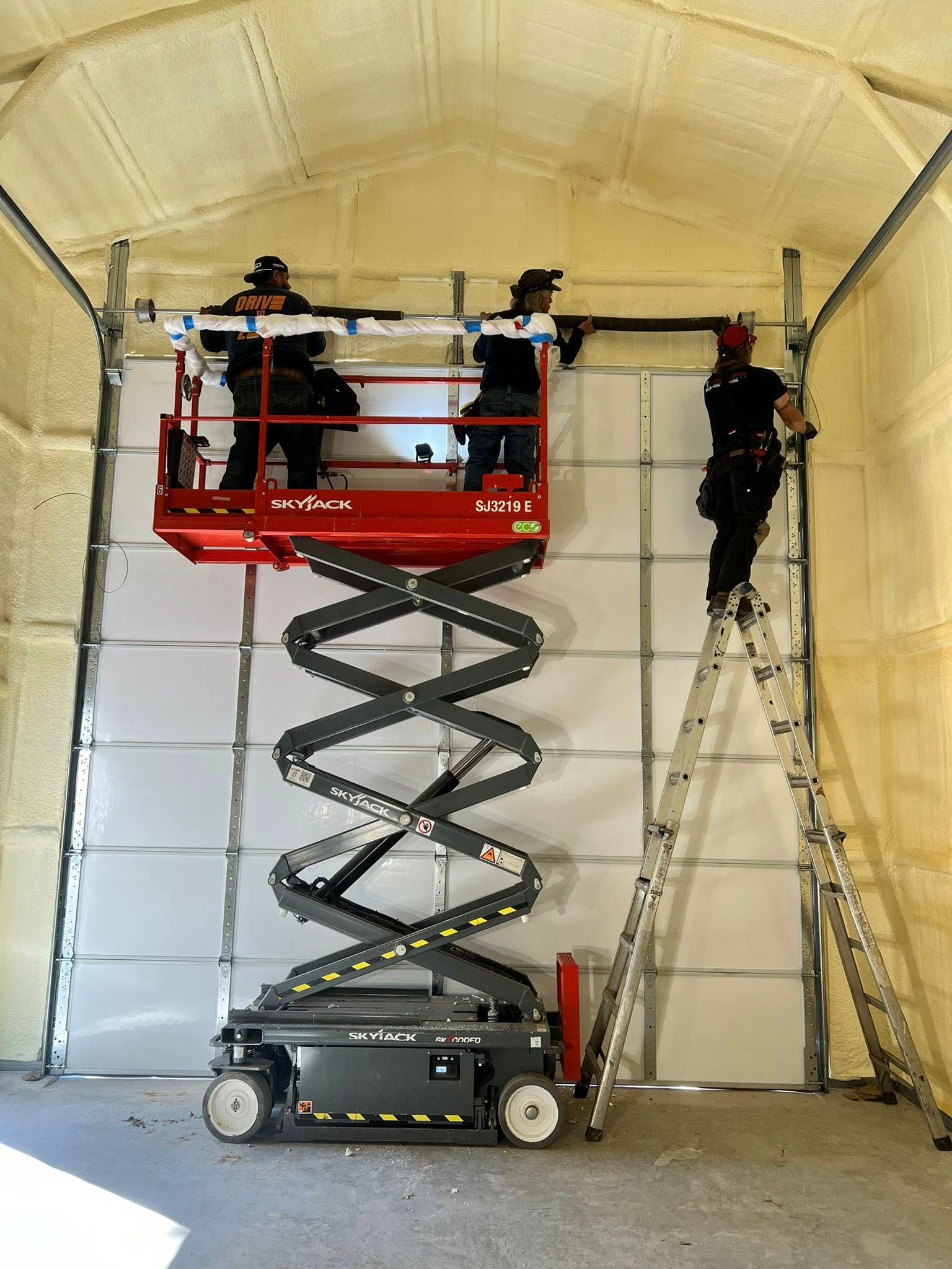 Workers installing a garage door on a lift and ladder in a building with spray foam insulation.