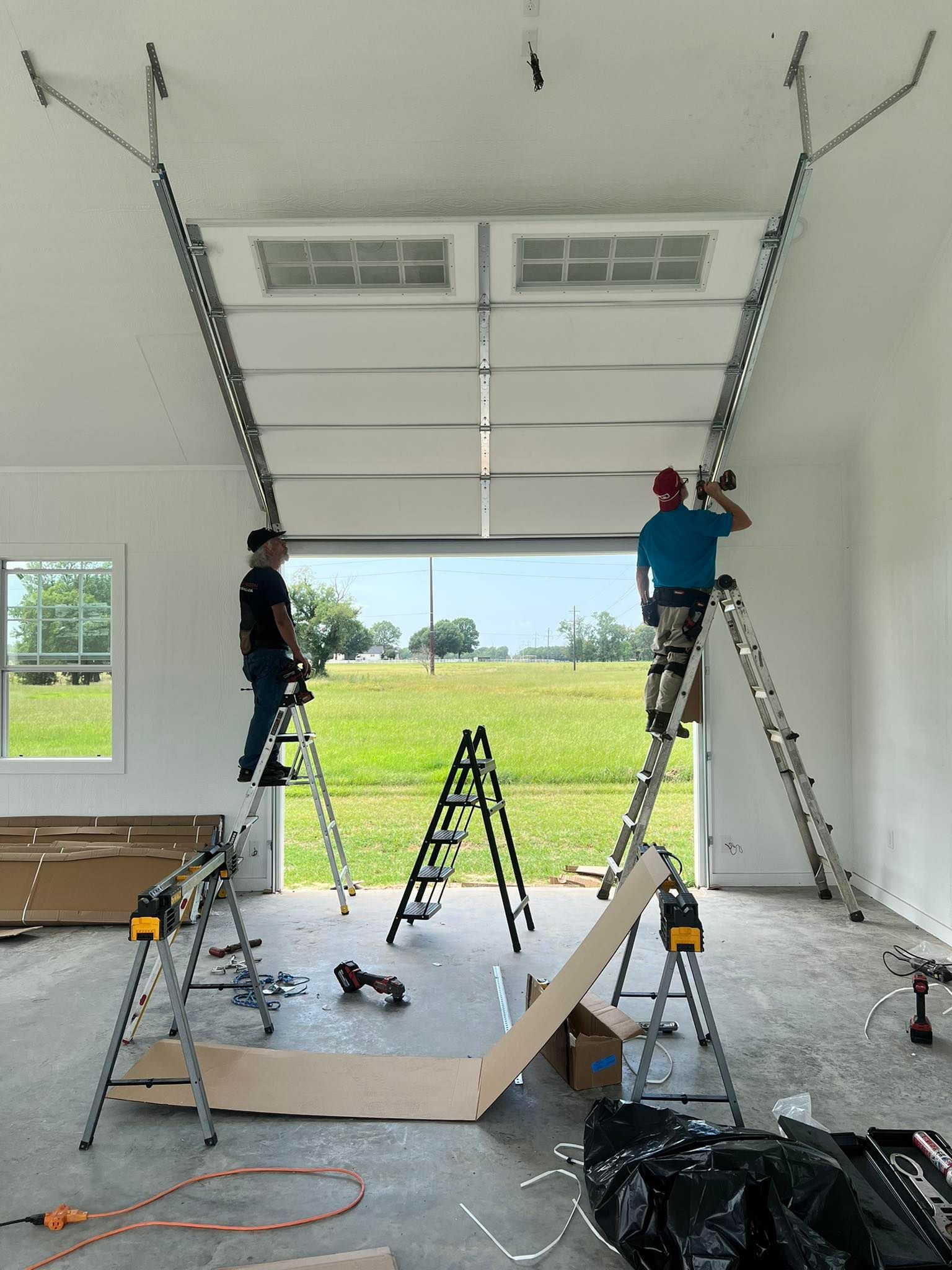 Two people installing a garage door on ladders inside a bright white garage, view to open field.