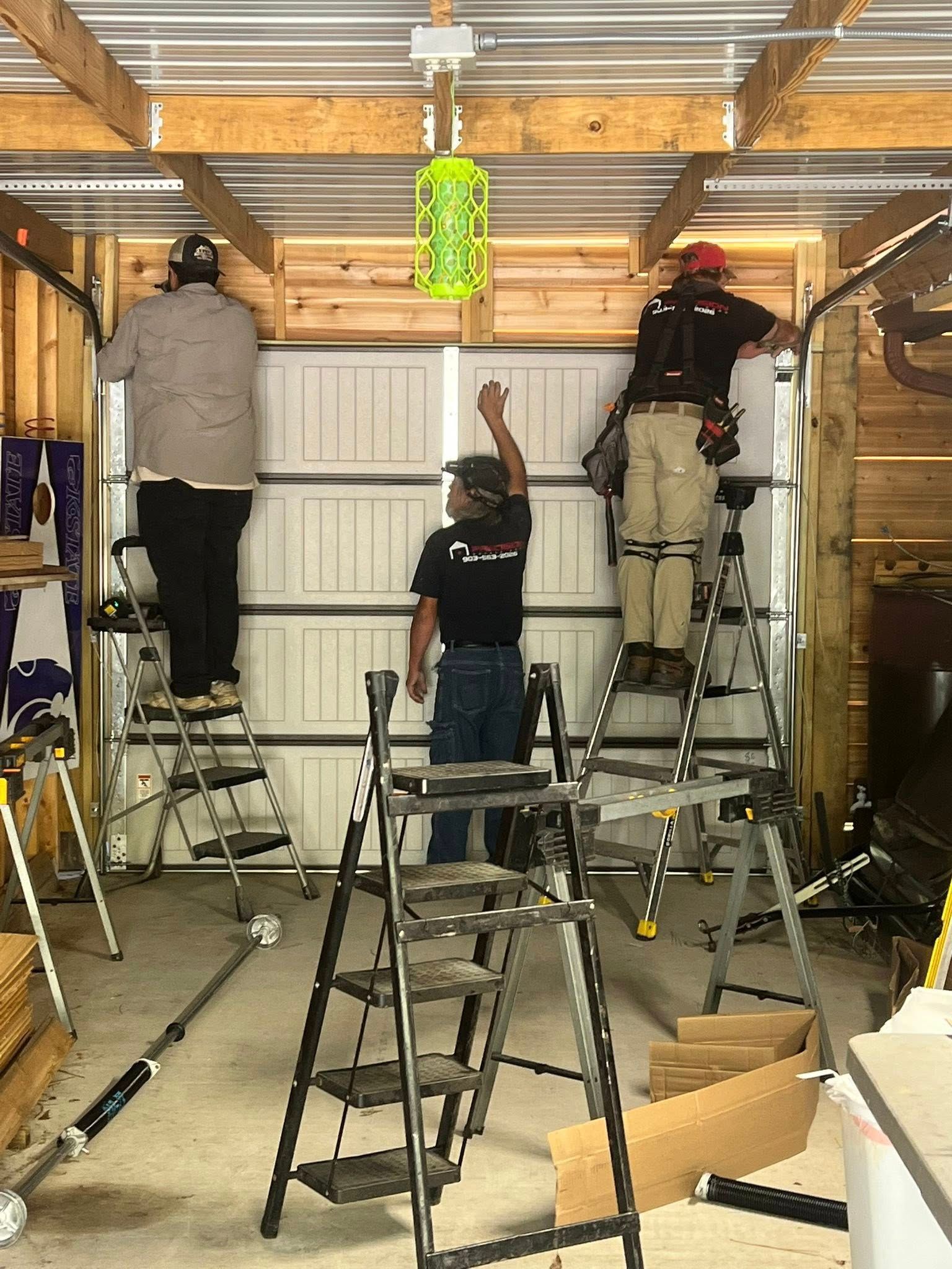 Three workers install a garage door, standing on ladders inside the garage.