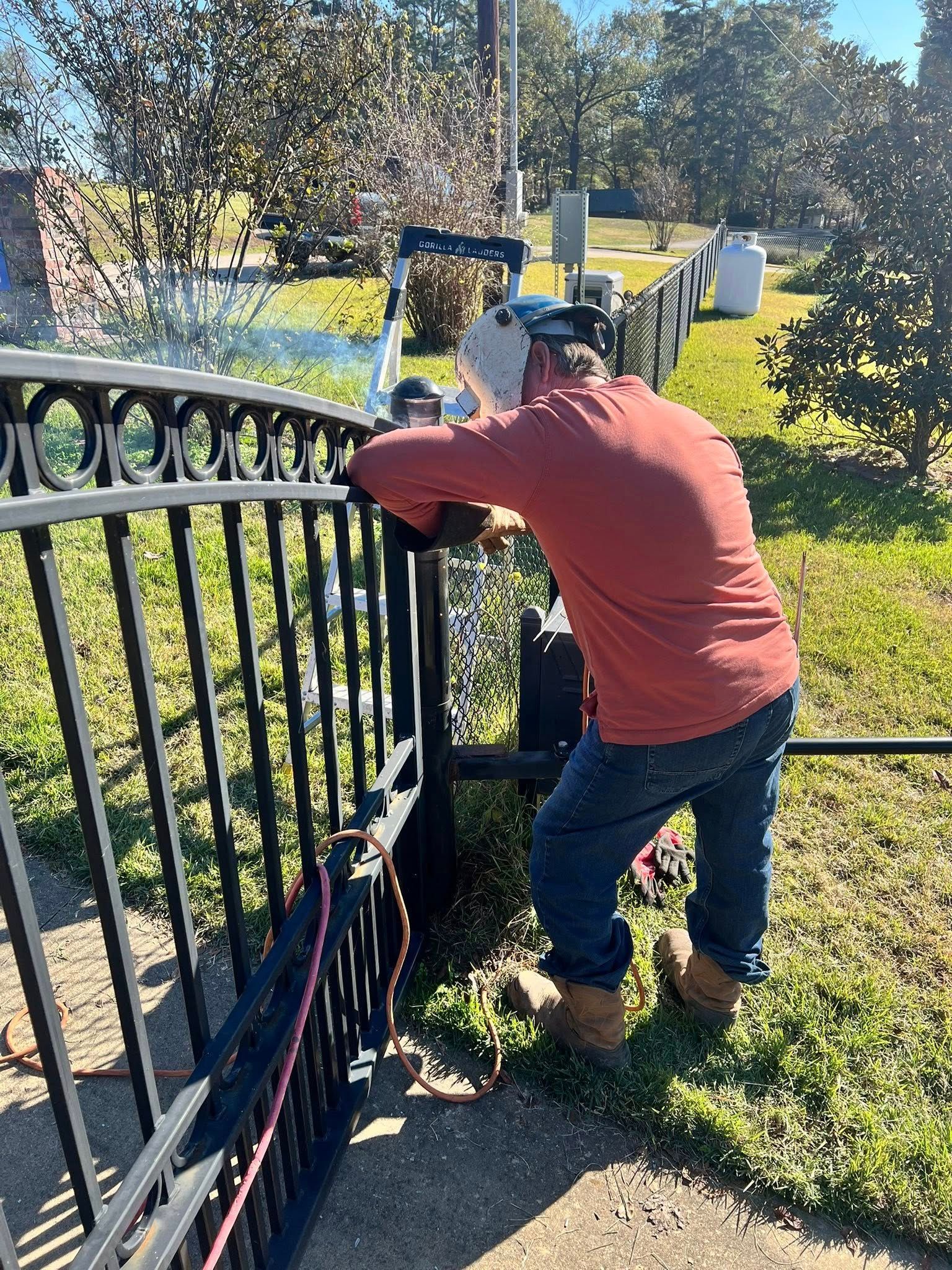 Man in red shirt working on a black metal gate outdoors; sunny day.