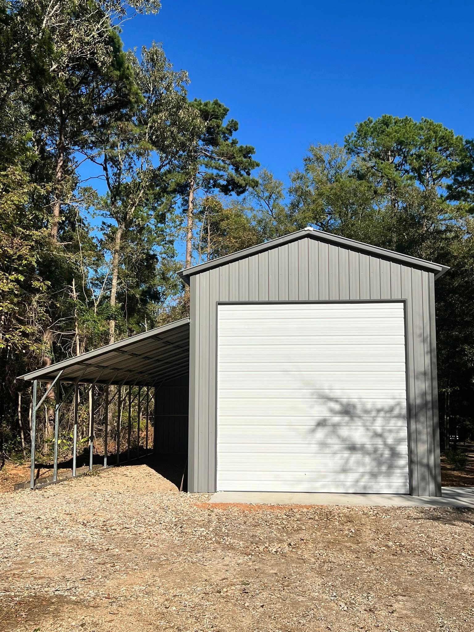 Gray metal building with a white garage door, carport, and gravel driveway, set in a wooded area under a blue sky.
