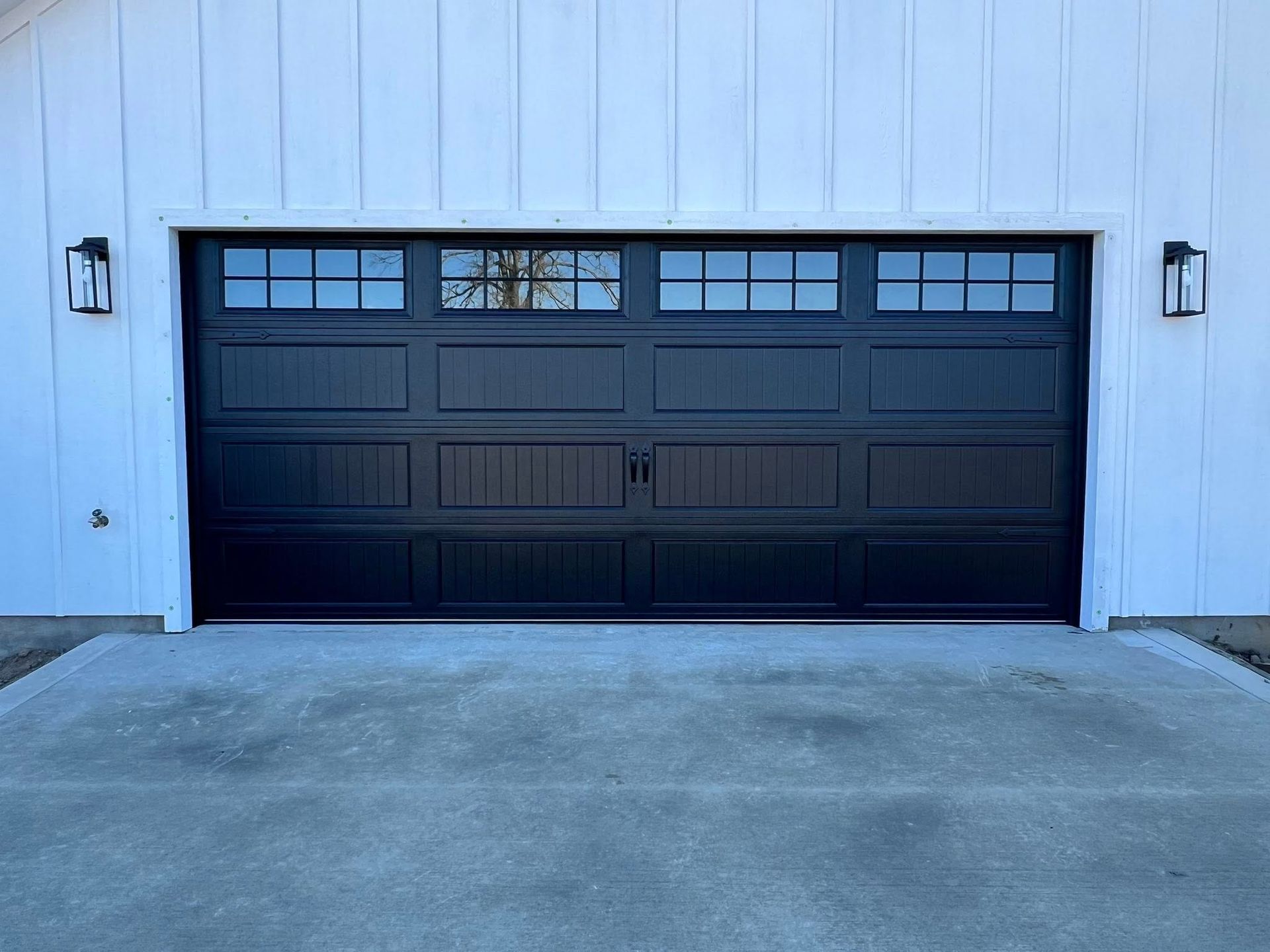 Black garage door with upper window panels, set in a white building with exterior lights. Concrete driveway.