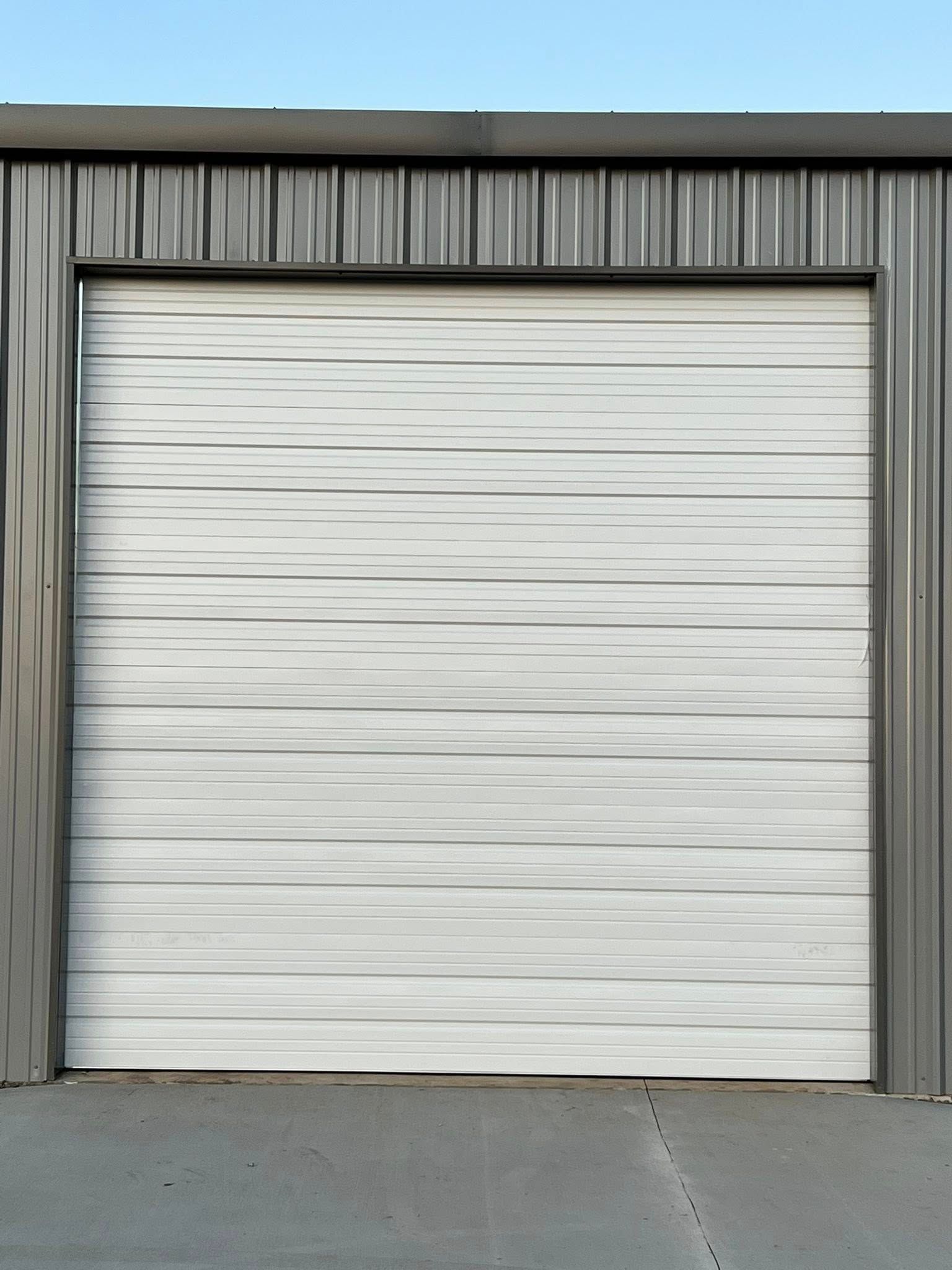 White garage door in a gray metal building, concrete ground, blue sky.