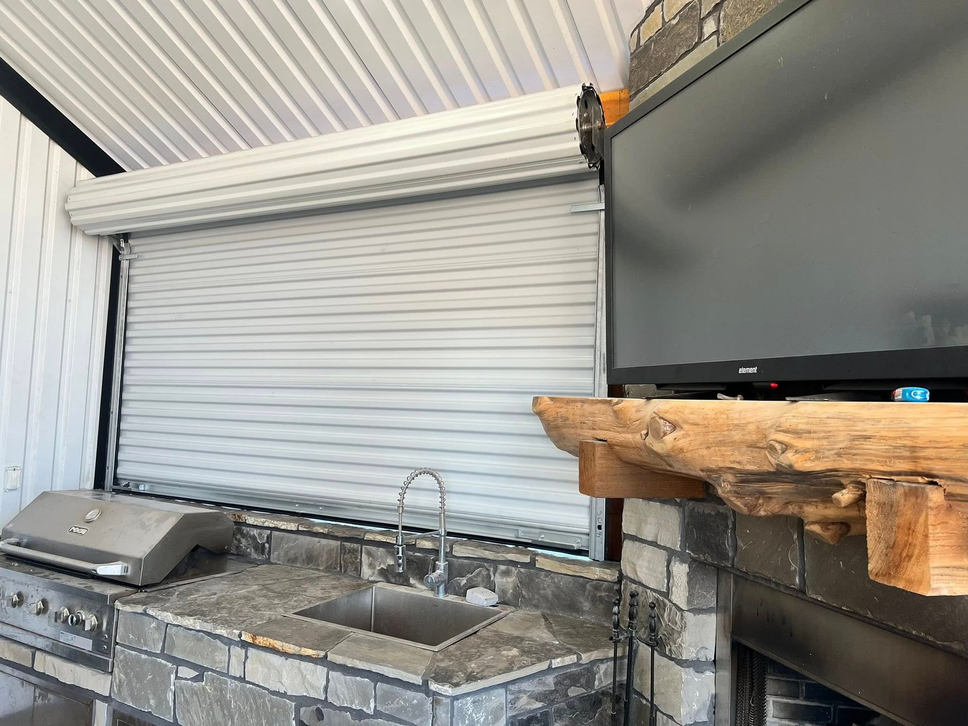 Outdoor kitchen with a closed roll-up shutter, grill, sink, and TV above a fireplace.