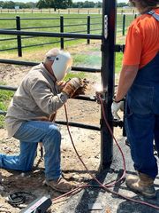 Man welding metal fence, kneeling, wearing welding mask, gloves, and jeans. Another person standing nearby.