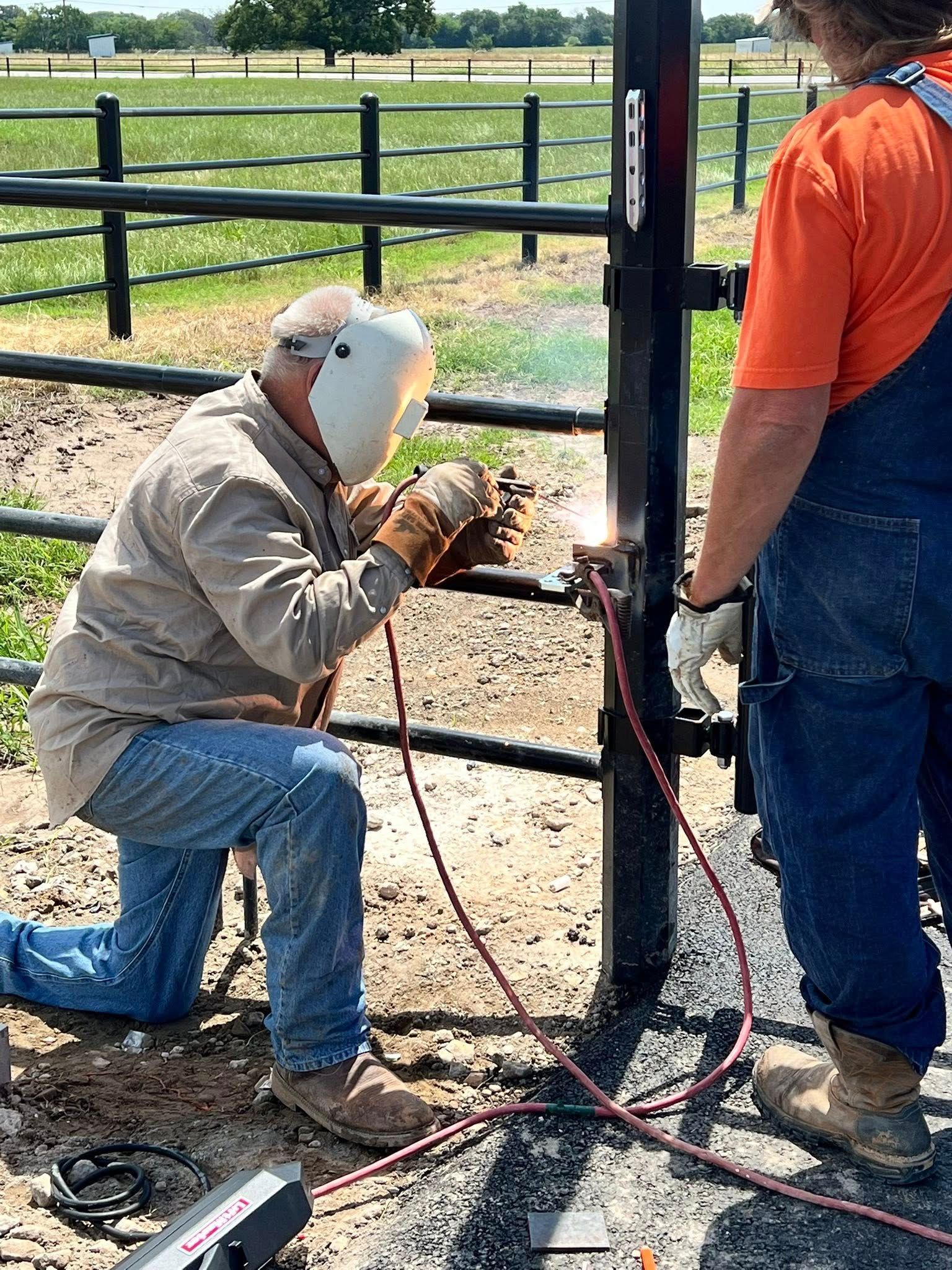 Man welding metal gate post while another watches. Outdoor setting with green grass and fence.