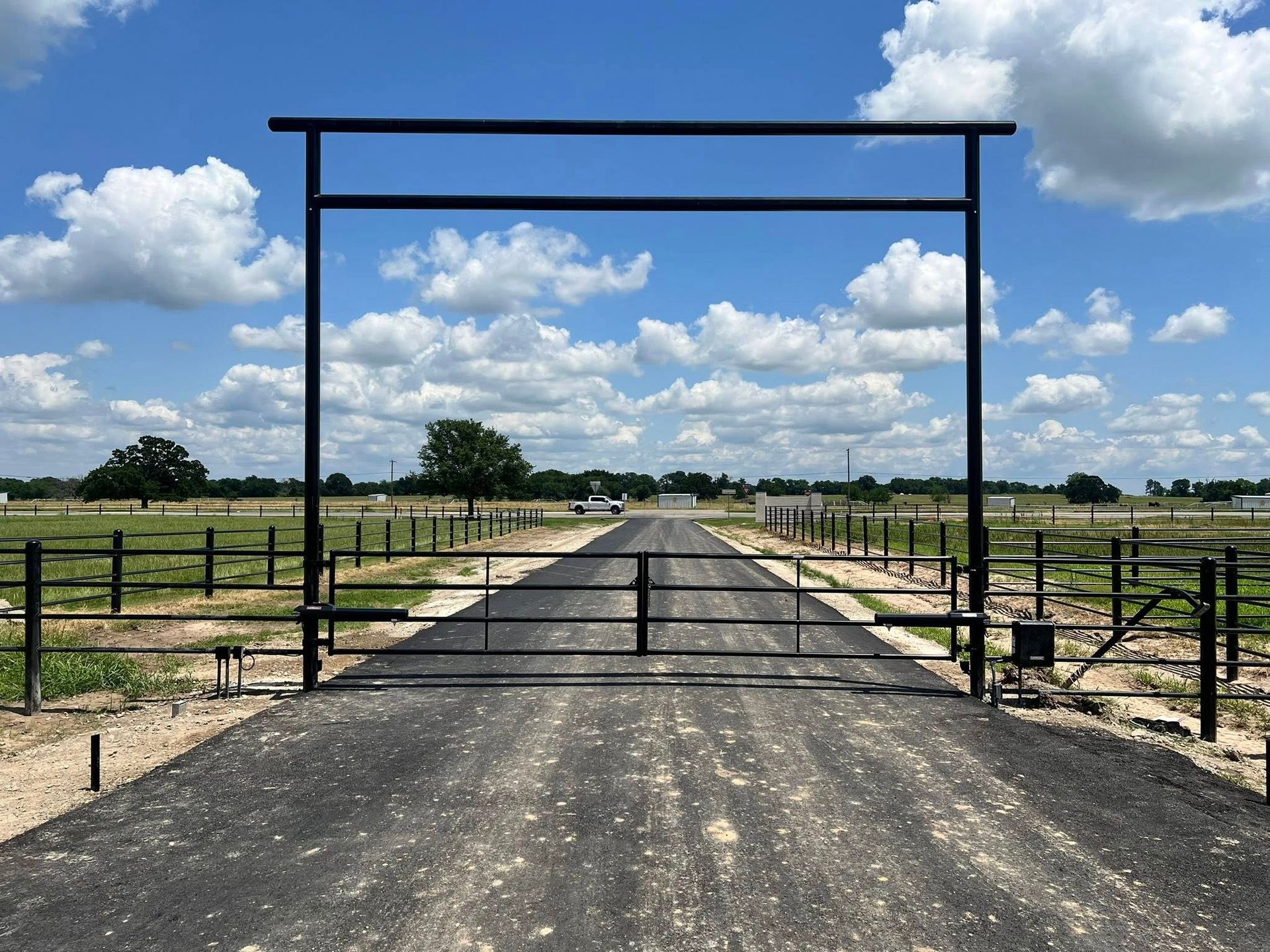 Black gate opens onto a paved driveway leading into a rural field under a blue sky with clouds.