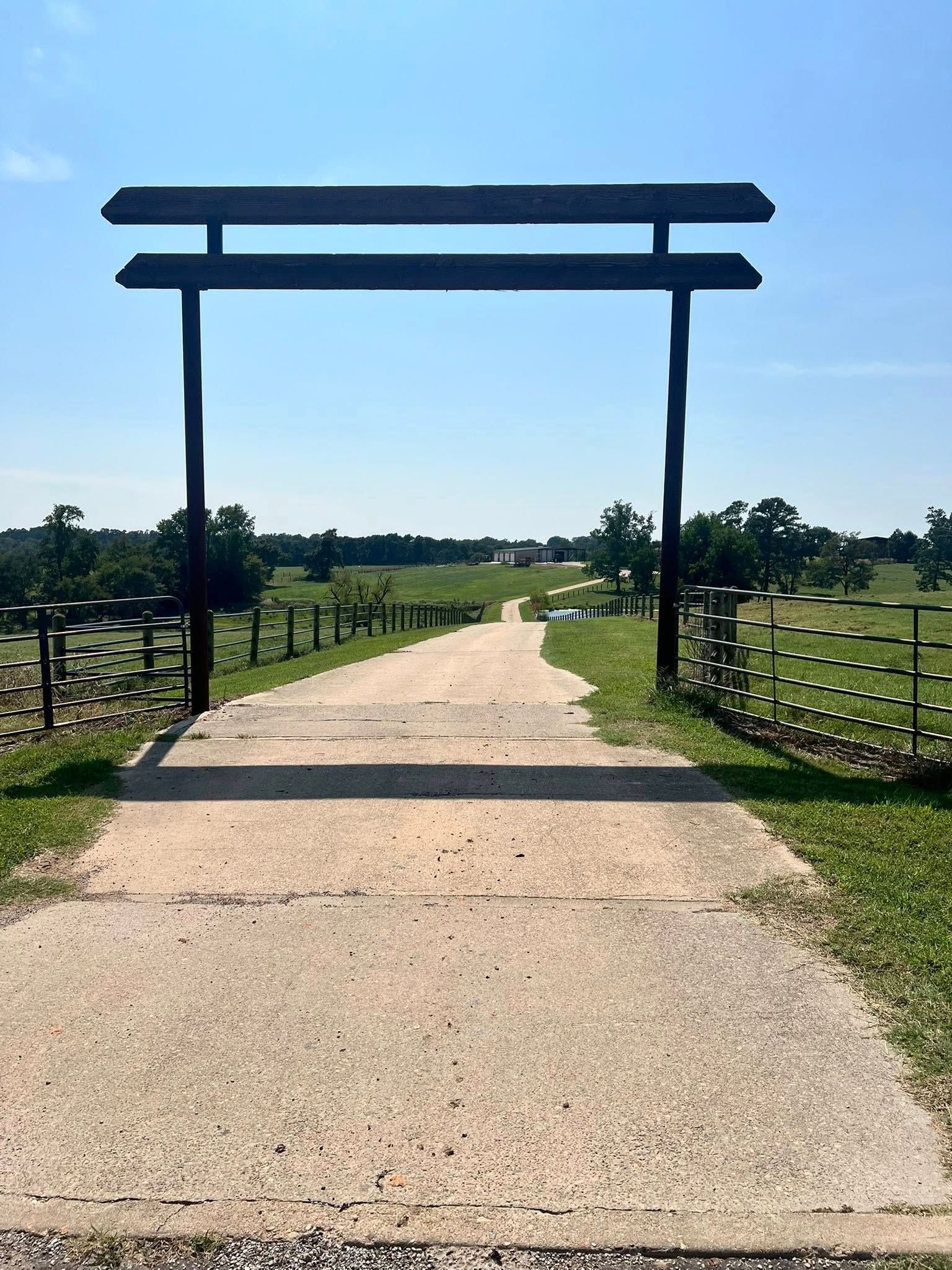 Concrete path leads to a gate with an overhead beam, and open field with trees in the distance.