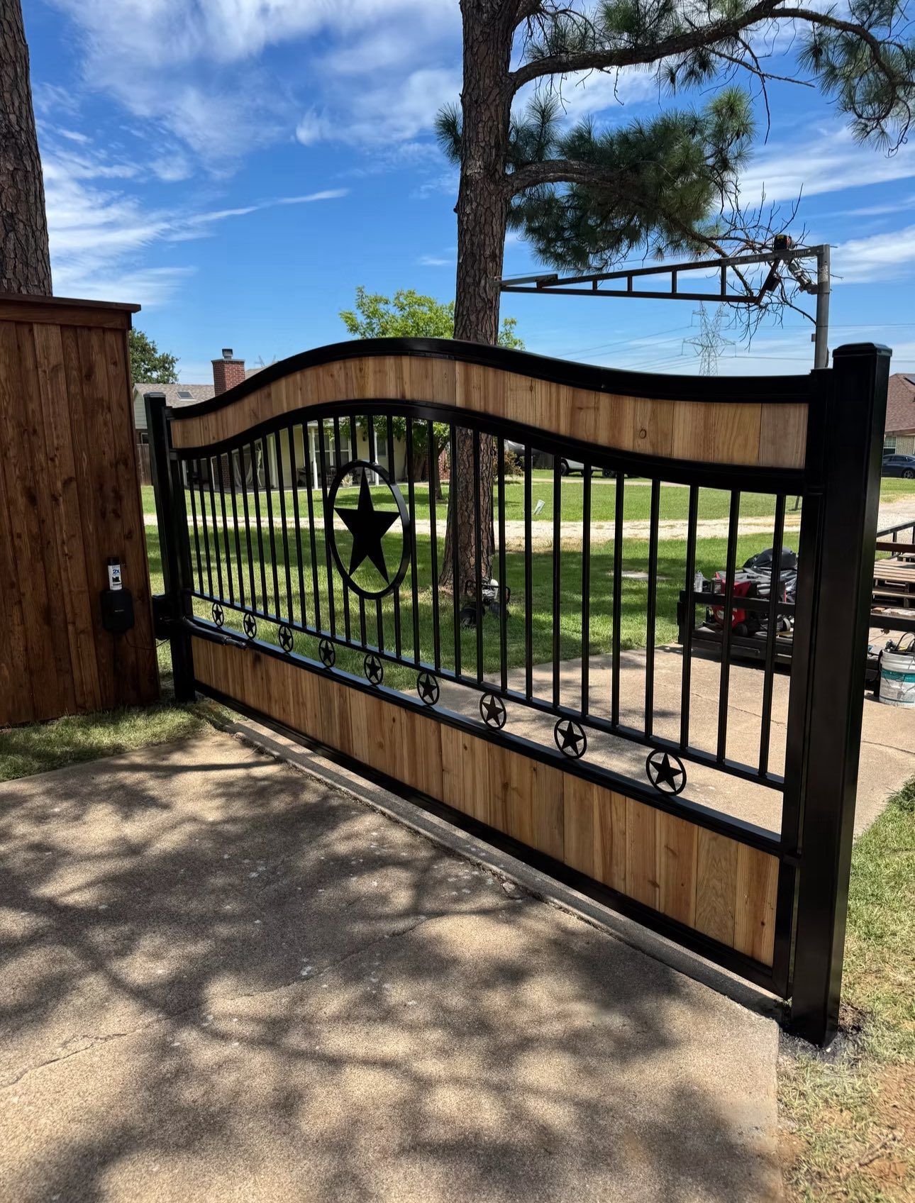 Wooden and black metal driveway gate with a star emblem, set against a sunny sky.