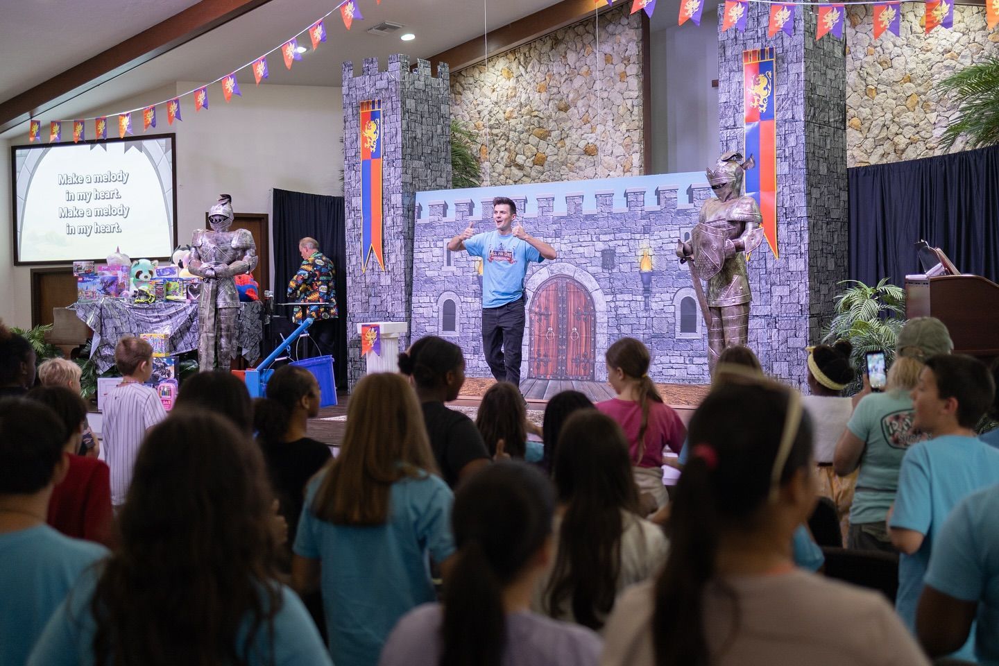 A man gestures on a stage with castle backdrop, knights, and an audience of children.
