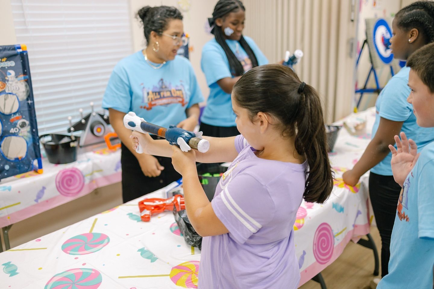 Girl aiming a toy gun at a game table. Several people watch, inside, bright colors, birthday setting.