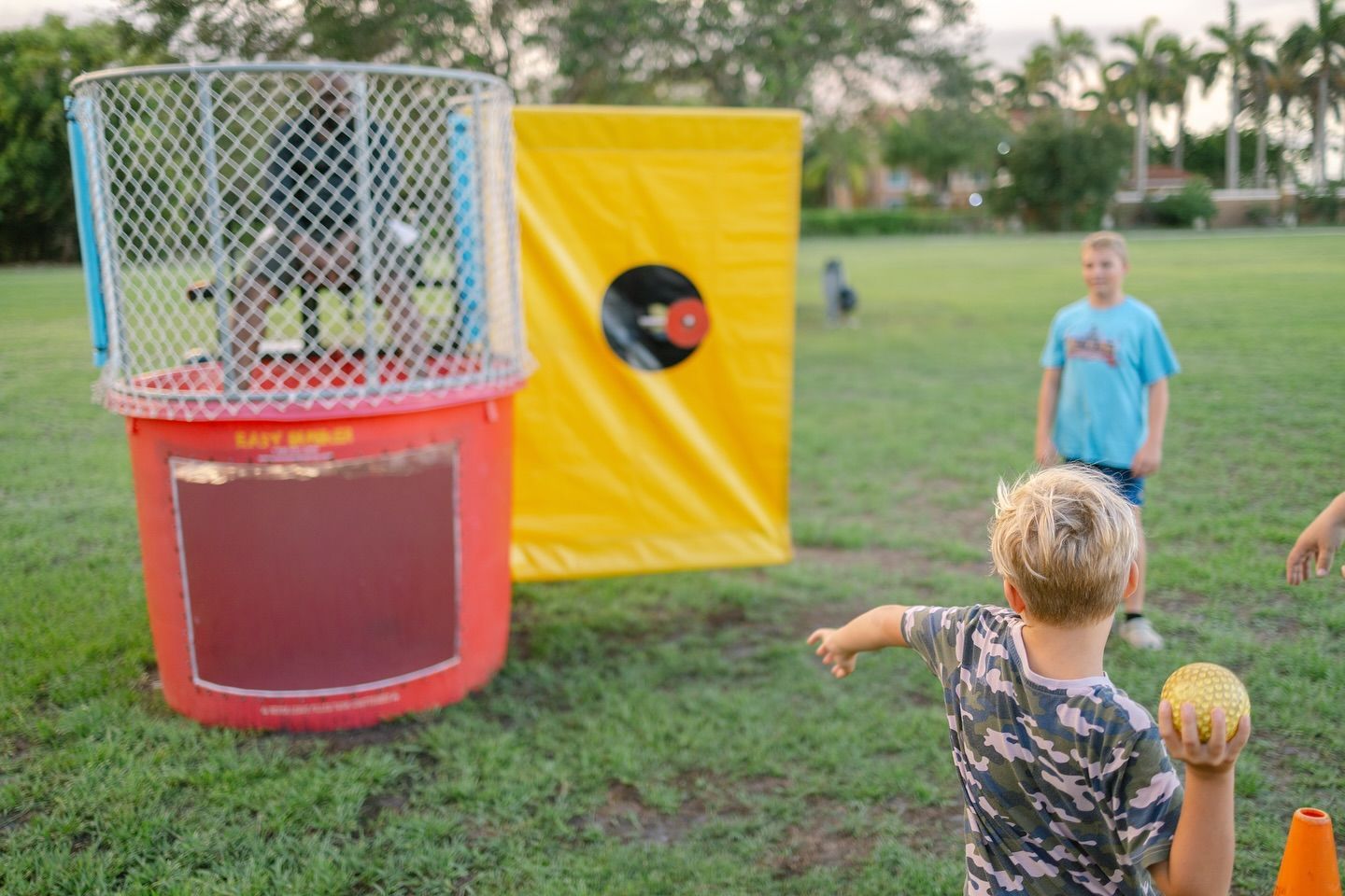 Boy about to throw a ball at a dunk tank, yellow target in background; outdoors on grass.