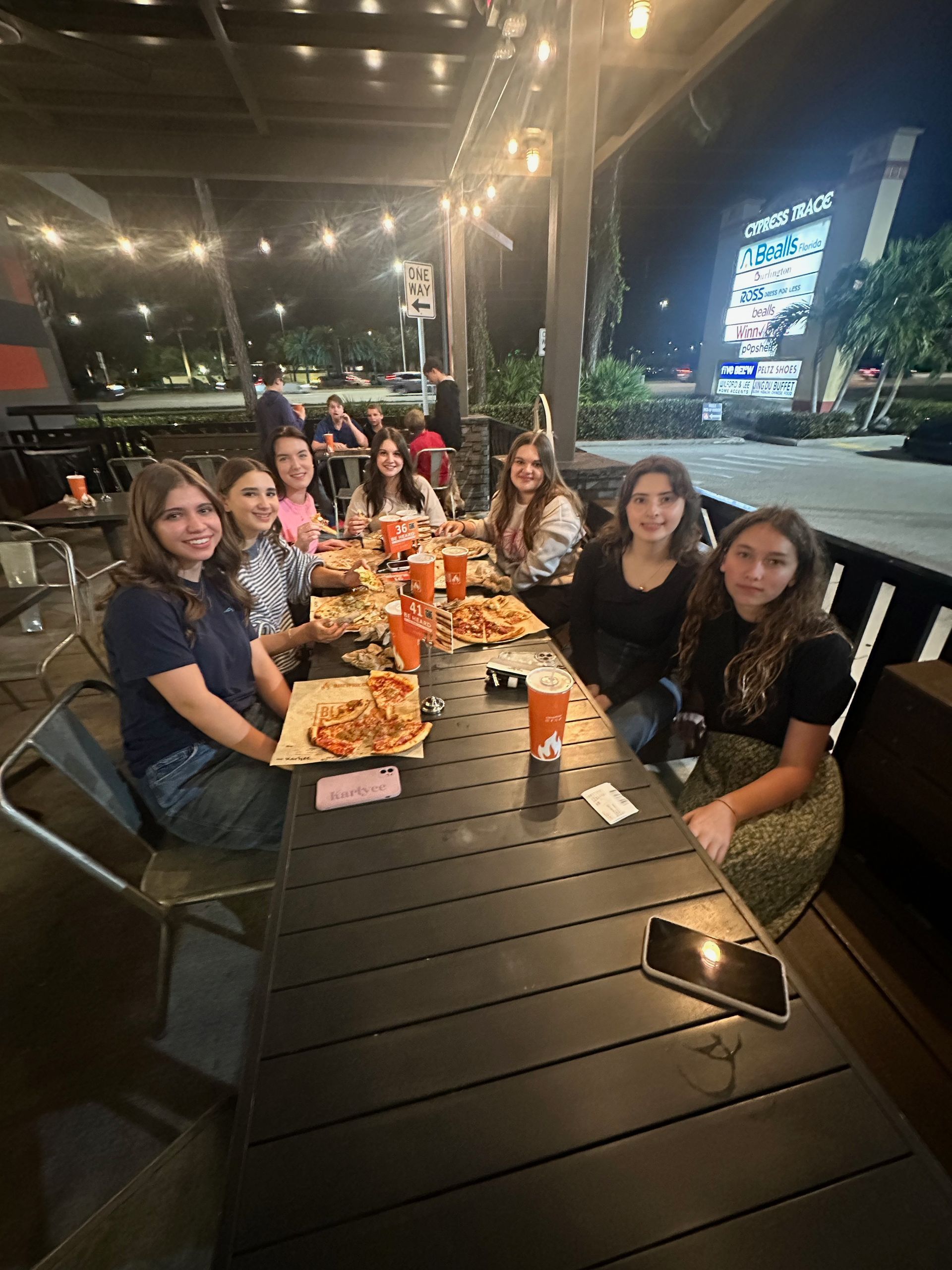 Group of friends smiling at a table, outside at night. Pizza, drinks. Lights overhead, First Bay sign in background.