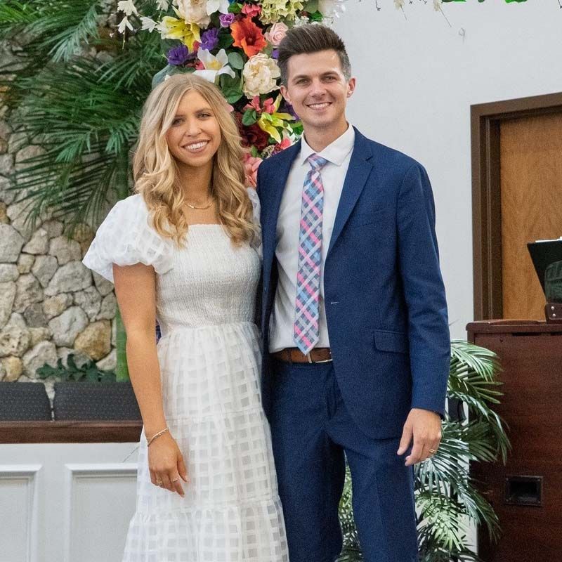 Couple in a church, woman in white dress, man in blue suit, smiling, standing in front of flowers.