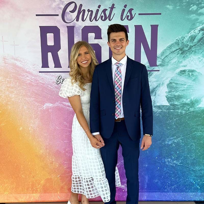 Couple in a church, woman in white dress, man in blue suit, smiling, standing in front of flowers.