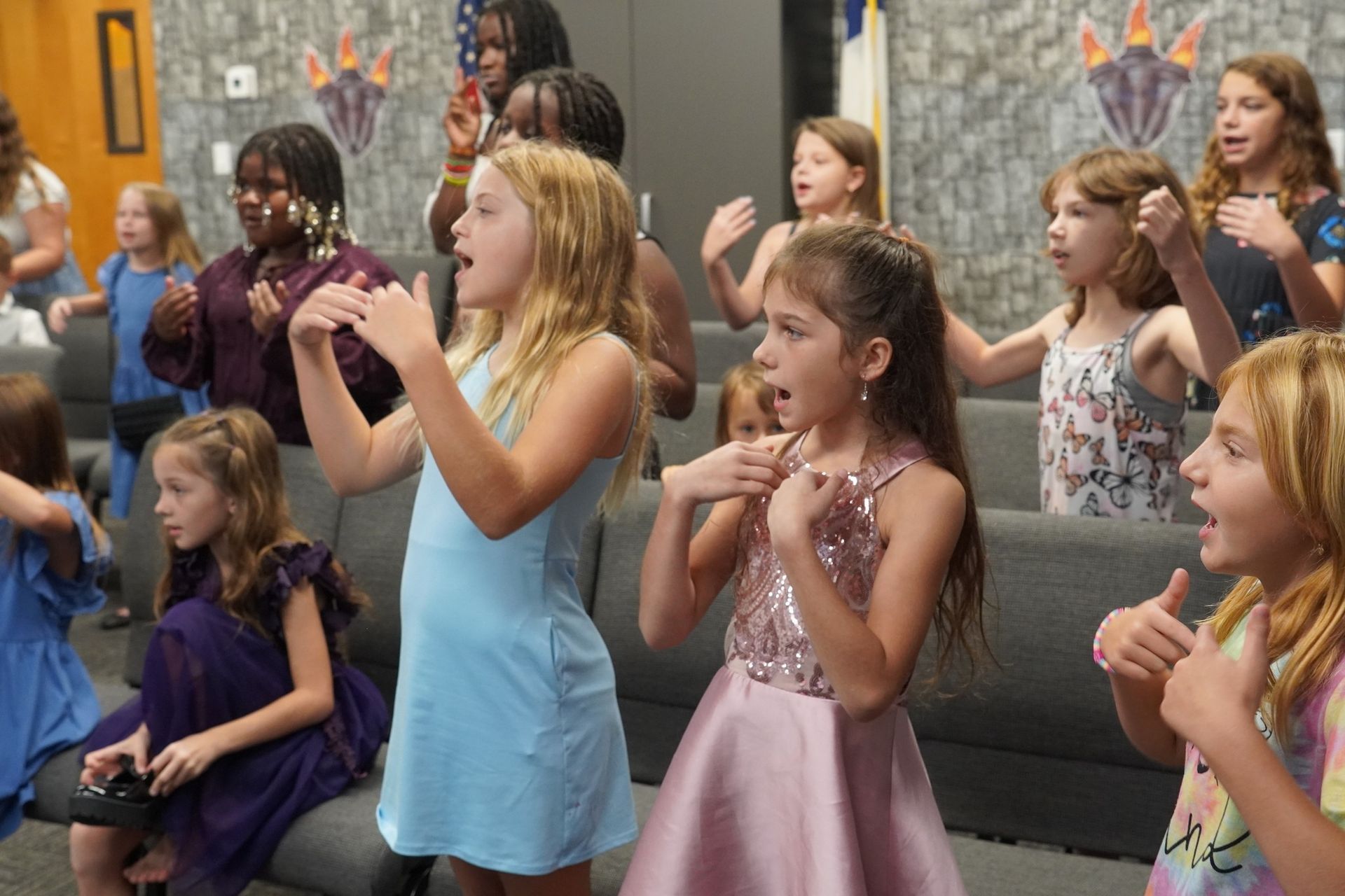 Group of young girls singing, some gesturing, in a room with patterned walls.