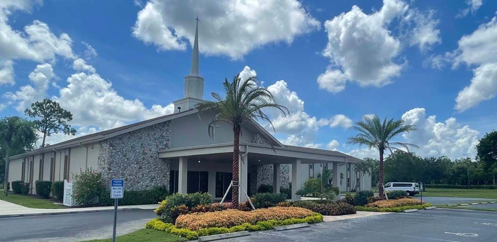 Church building with a steeple and palm trees against a cloudy blue sky.