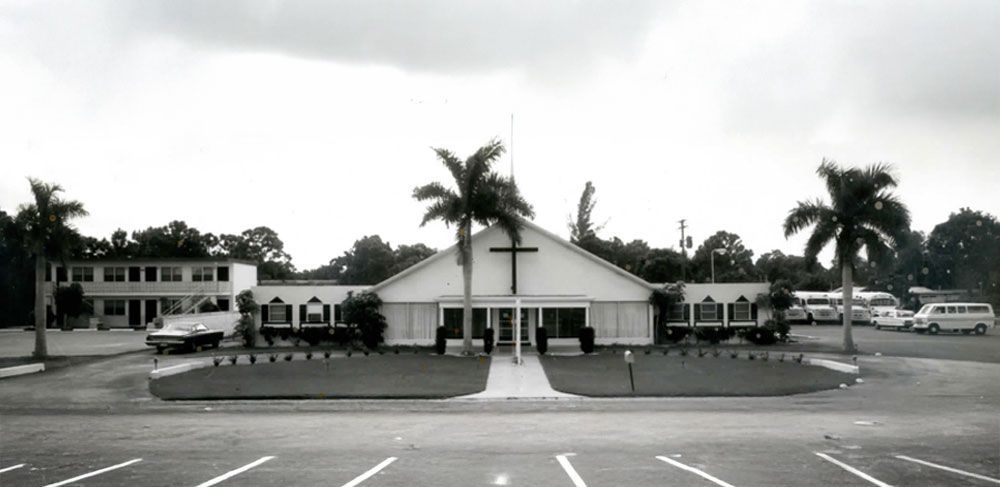 Black and white photo of a church with a large cross, flanked by palm trees, and a parking lot.