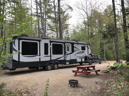 A rv is parked in a gravel lot in the woods next to a picnic table.