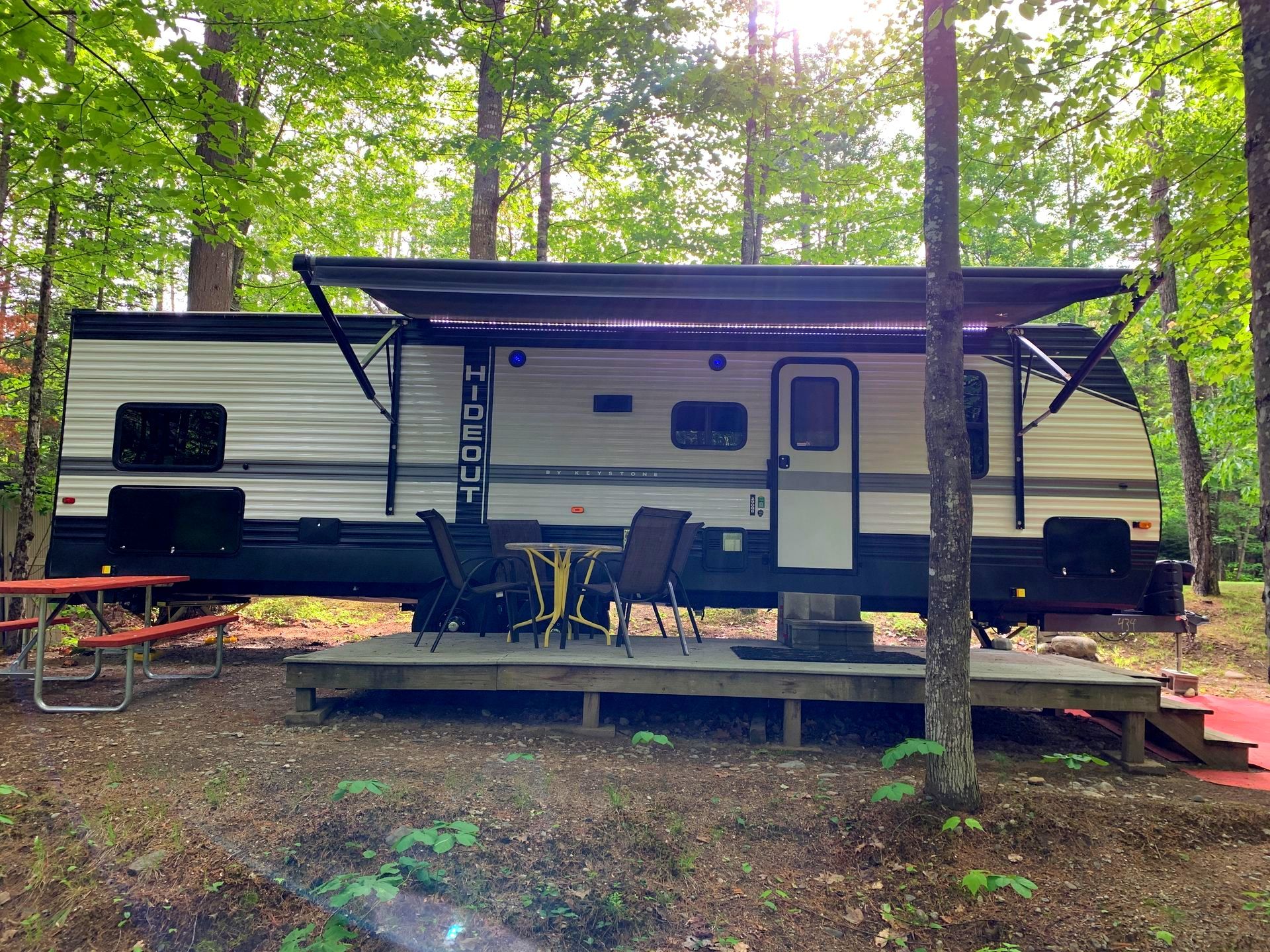 A rv is parked in the woods with a table and chairs on the deck.