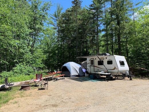 A rv and tent are parked in a gravel lot in the woods.
