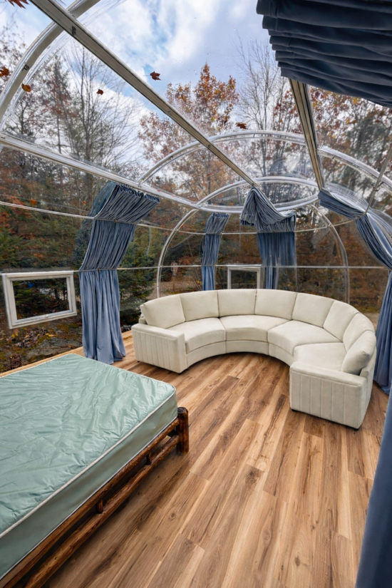Cozy glass sunroom with cream sectional sofa, wood floor, and bed beside blue curtains.