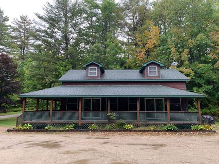Rustic log cabin with green roof and wraparound porch, set among trees on a wooded lot.