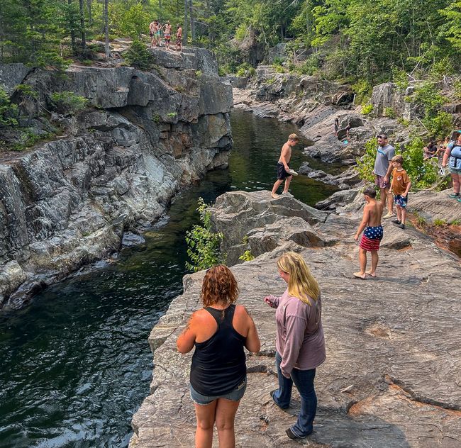 People standing on rocky ledges above a dark green swimming hole in a wooded gorge