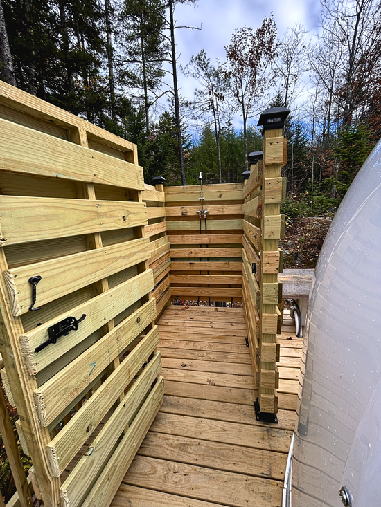 Wooden outdoor walkway beside a white greenhouse, with slatted fence panels and trees in the background