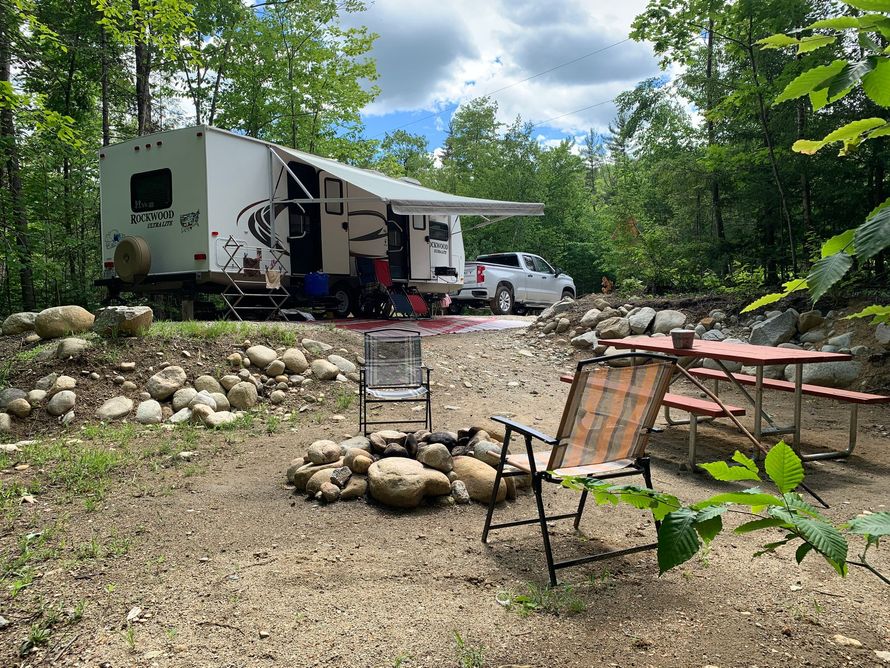 A rv is parked in the woods next to a picnic table and chairs.