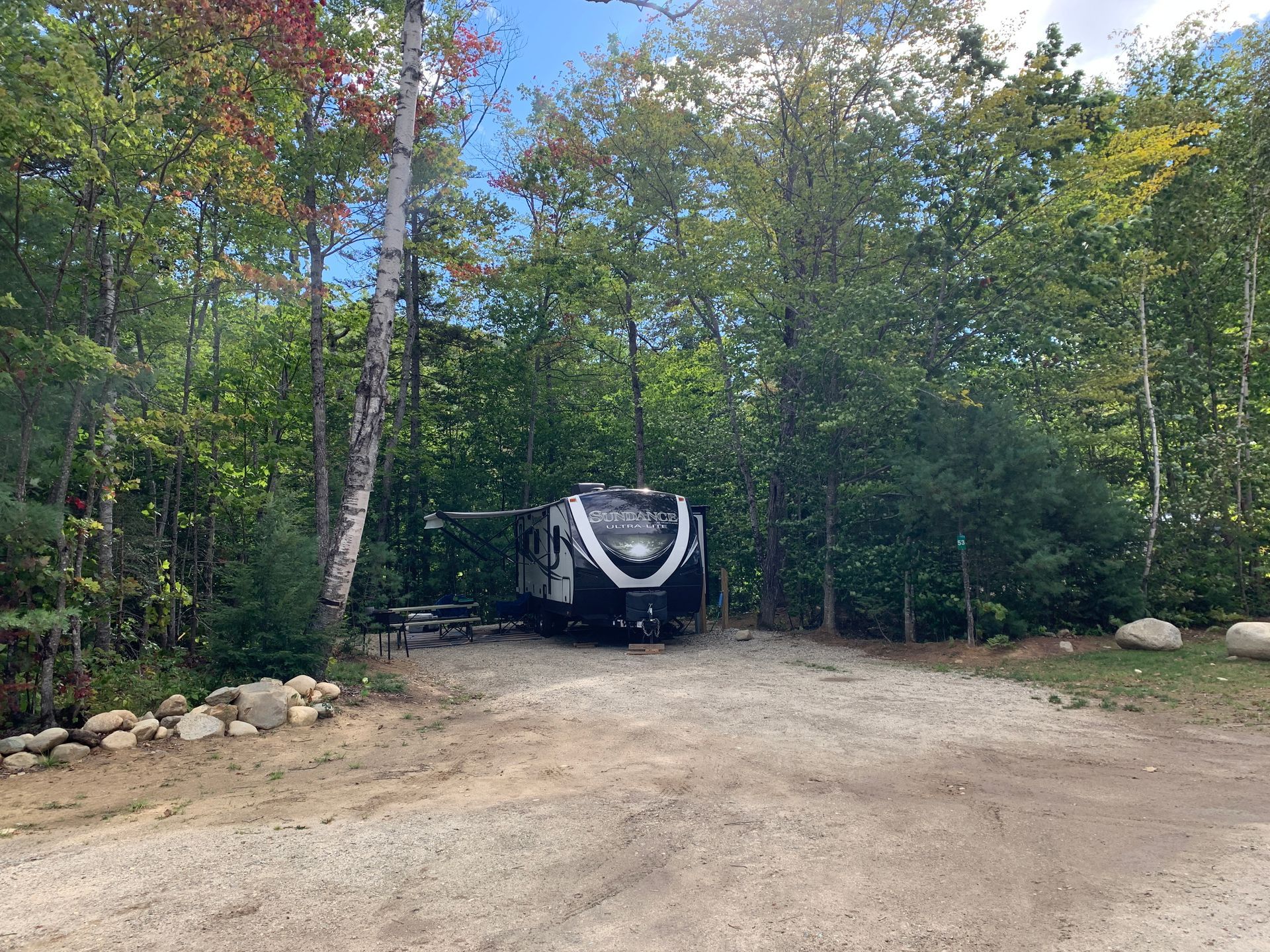 A rv is parked in a gravel lot in the middle of a forest.