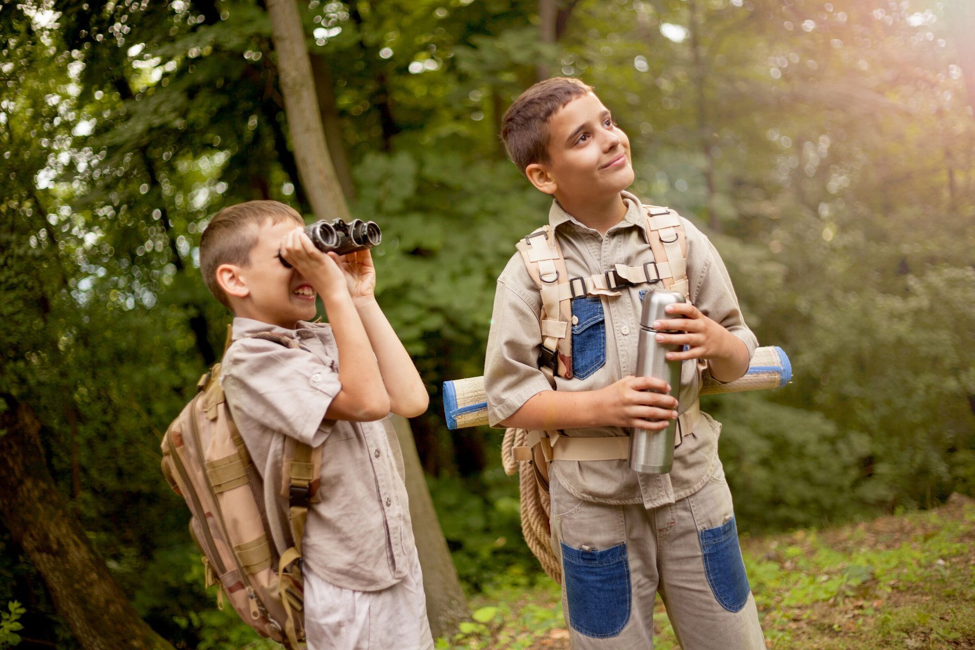 Two young boys are looking through binoculars in the woods.