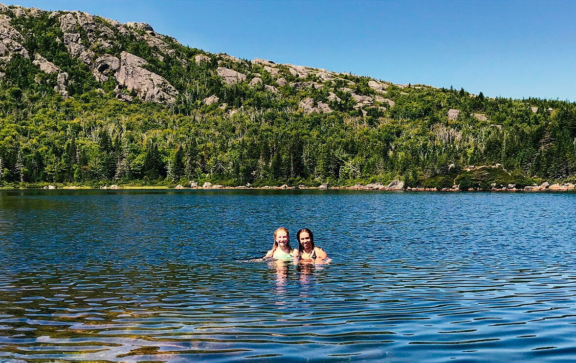 Two people are swimming in a lake with mountains in the background.