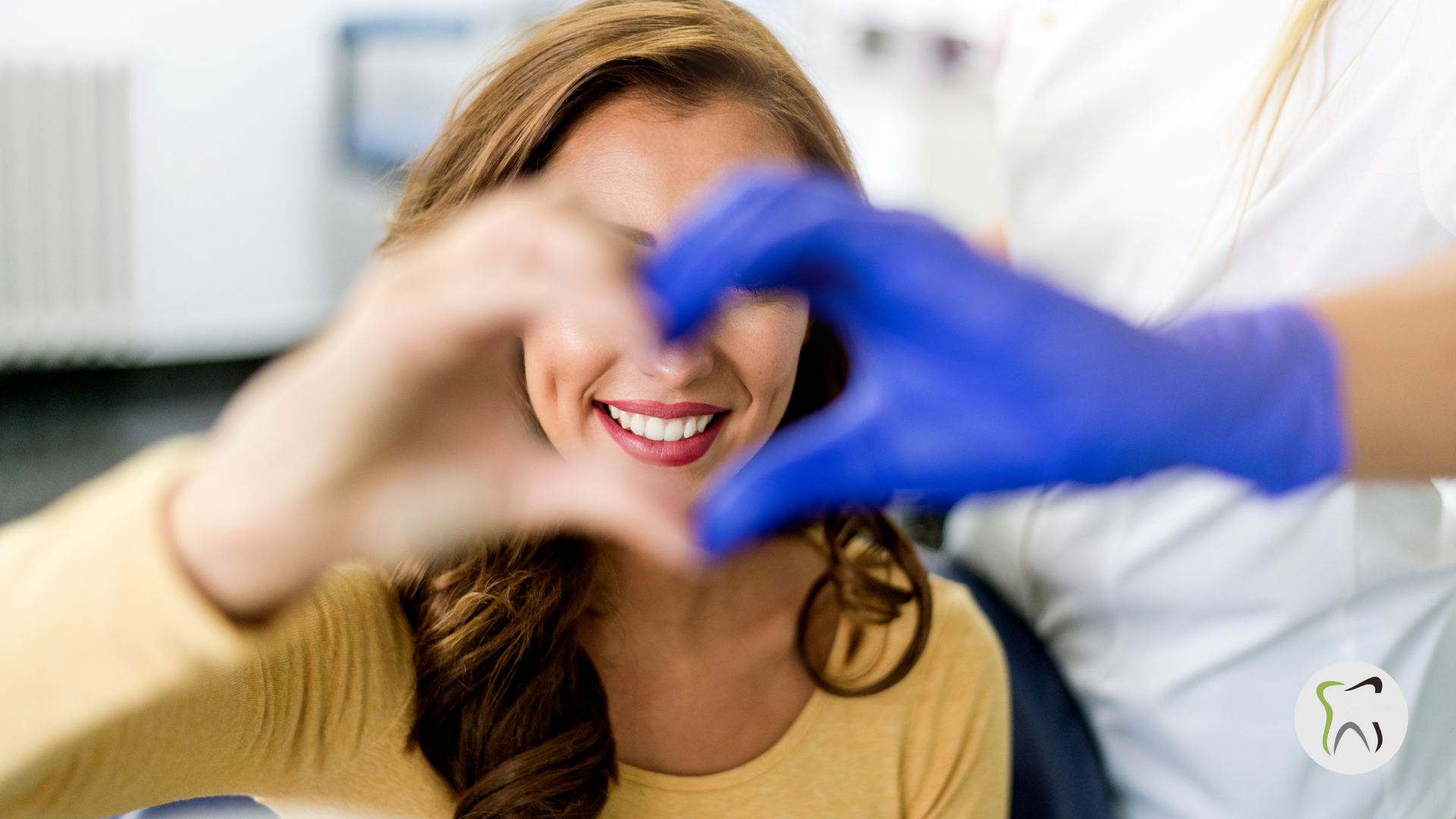 A woman is making a heart shape with her hands while sitting in a dental chair.