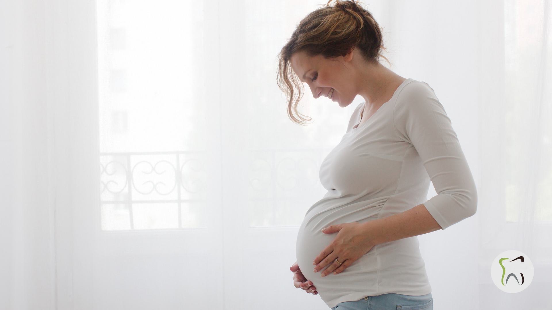 A pregnant woman is standing in front of a window holding her belly.