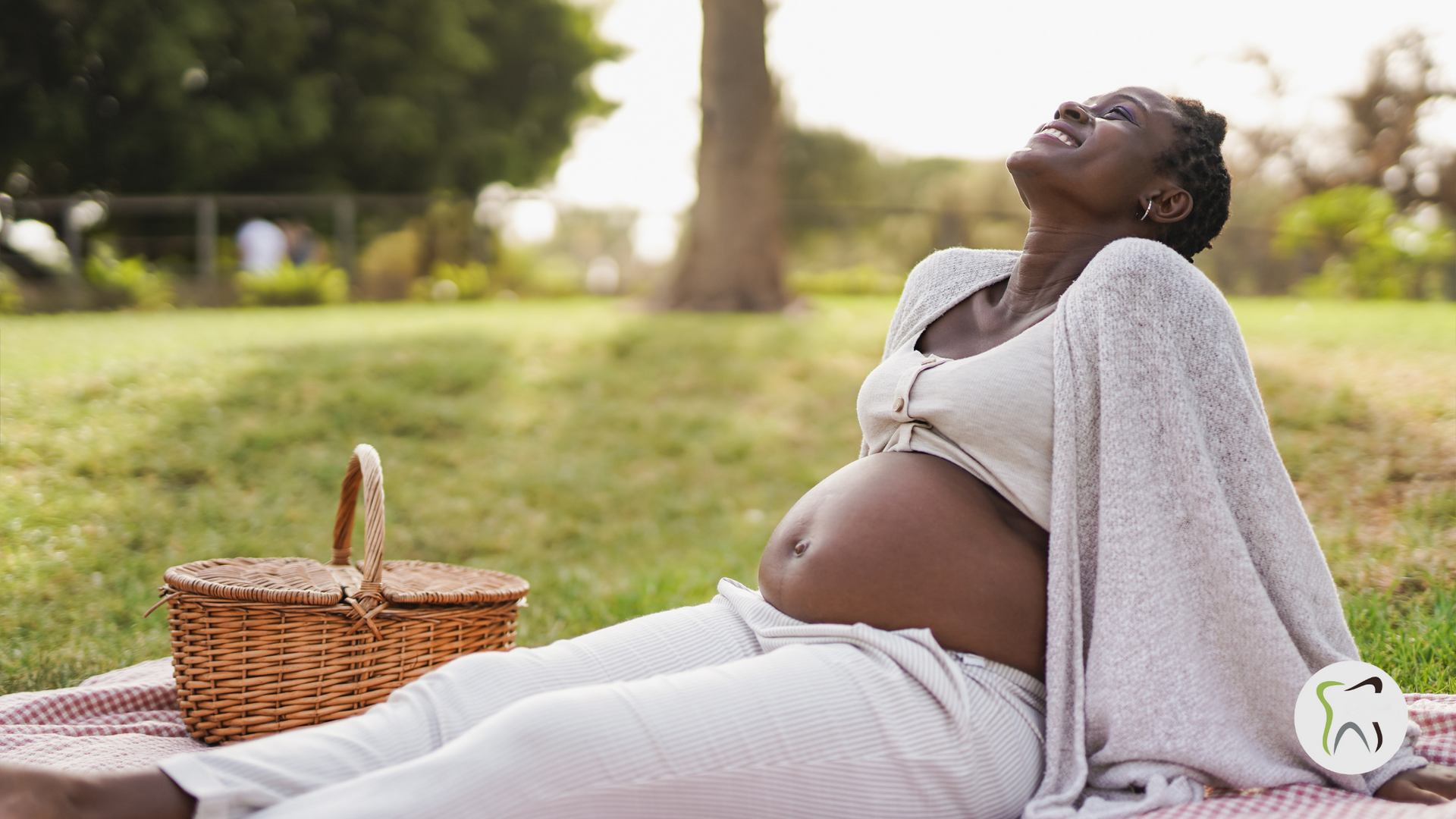 A pregnant woman is sitting on a picnic blanket in a park.