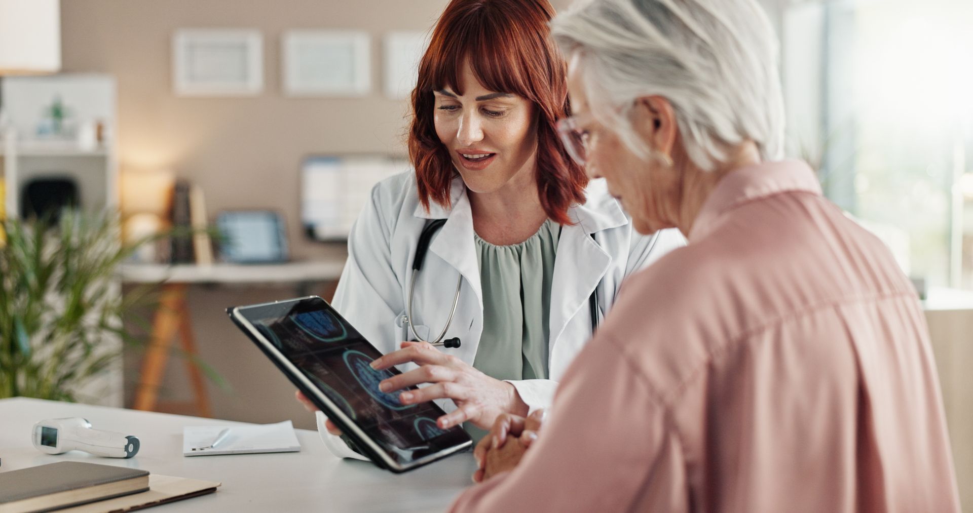 Doctor showing an older patient a tablet screen. They sit at a desk in a medical office.