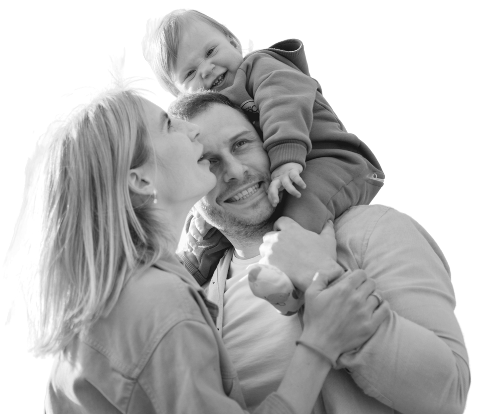 Family portrait: man carrying child on shoulders, woman kissing man's cheek, all smiling.