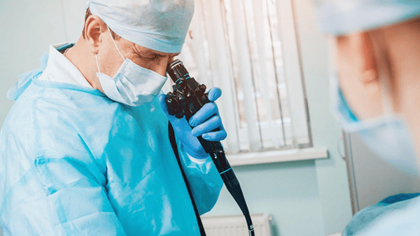Doctor in blue scrubs looks through an endoscope, another person visible nearby in a clinical setting.