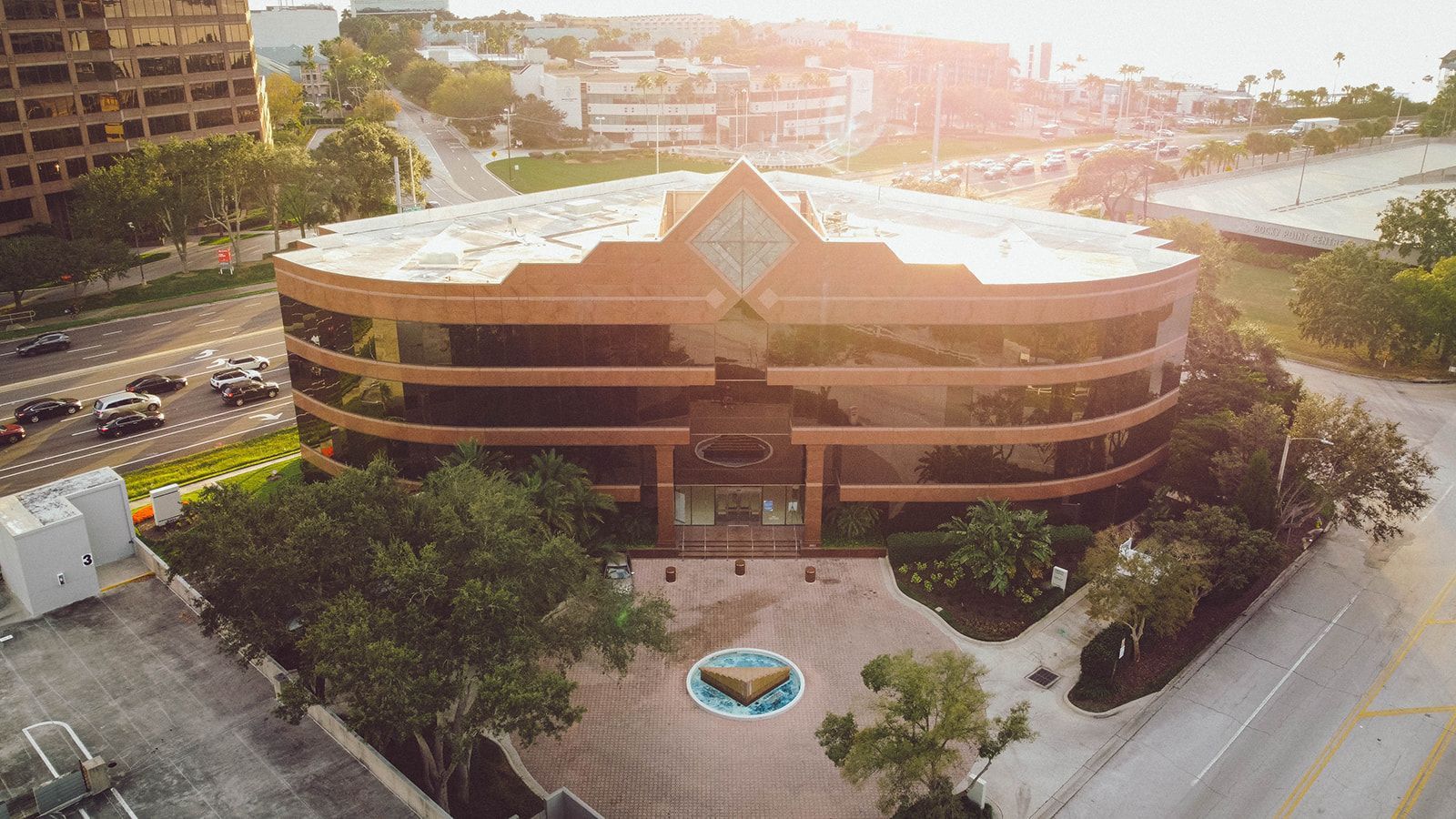 Brown building with glass windows, fountain, trees, and cars on road in front. Bright sunlight. Brown building with glass windows, fountain, trees, and cars on road in front. Bright sunlight.