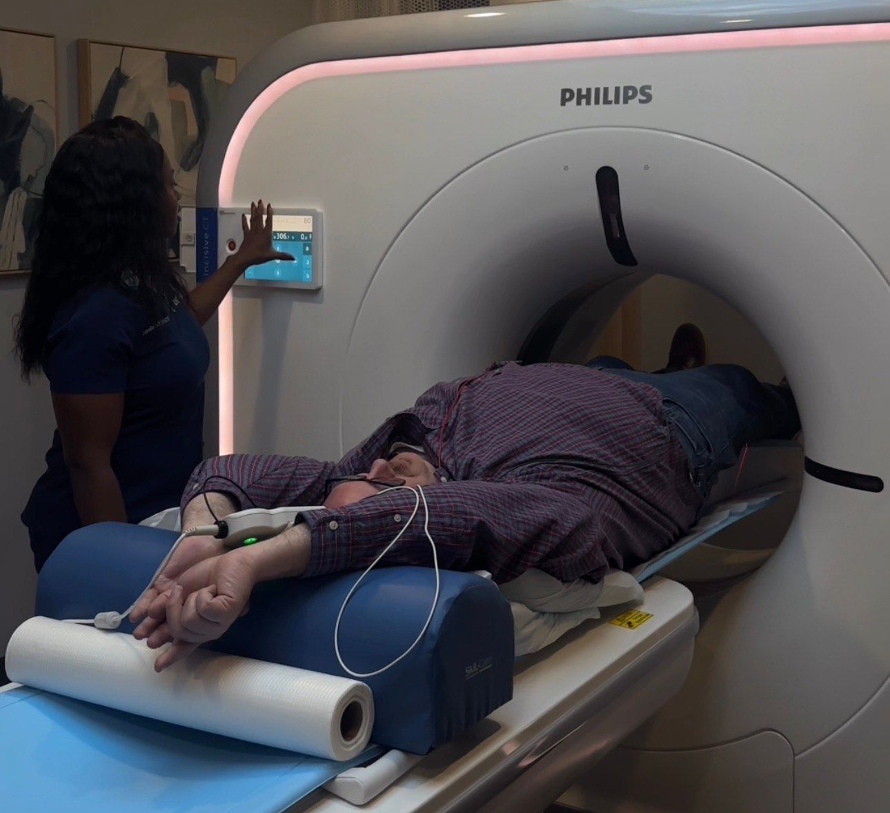 Woman in a blue gown undergoing a mammogram in a medical setting. A healthcare worker is in the background.