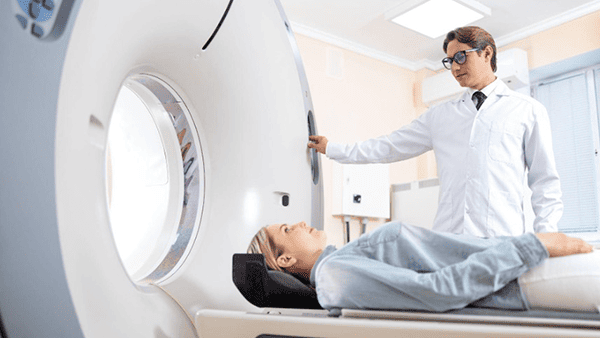 Woman lying in a CT scanner; doctor in white coat stands nearby, observing.