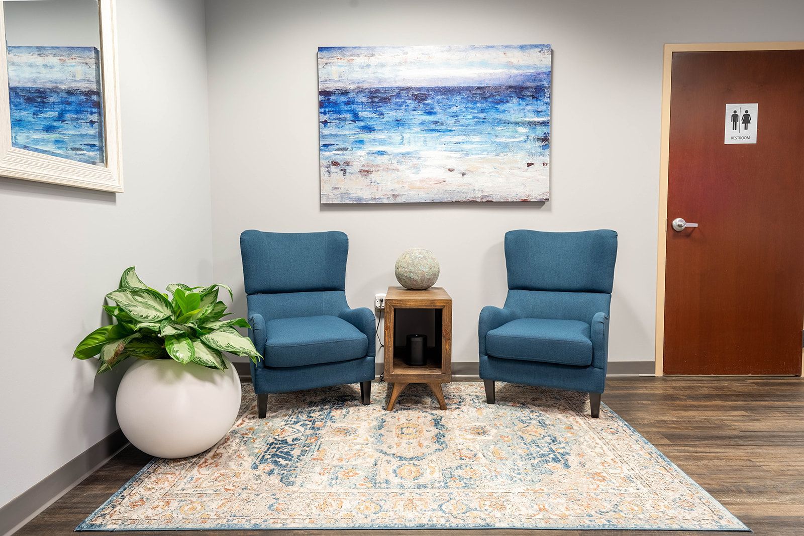 Two blue armchairs in waiting area with art and rug; a plant in a white pot, and a brown door. Two blue armchairs in waiting area with art and rug; a plant in a white pot, and a brown door.