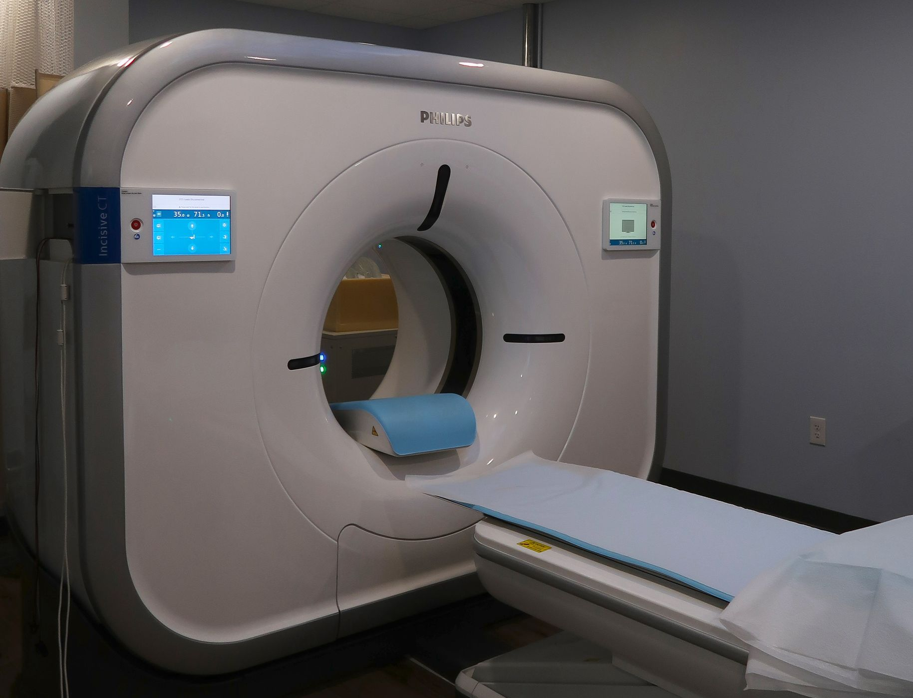 CT scan machine in a medical examination room, with a patient bed and storage cabinets. CT scan machine in a medical examination room, with a patient bed and storage cabinets.